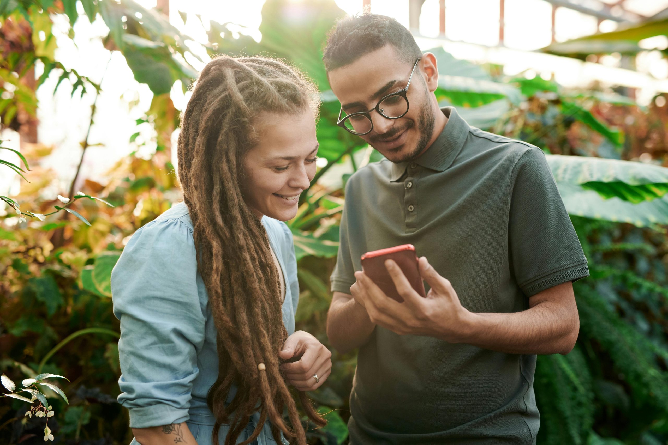 Two young adults smiling while using a smartphone in a lush green outdoor setting.